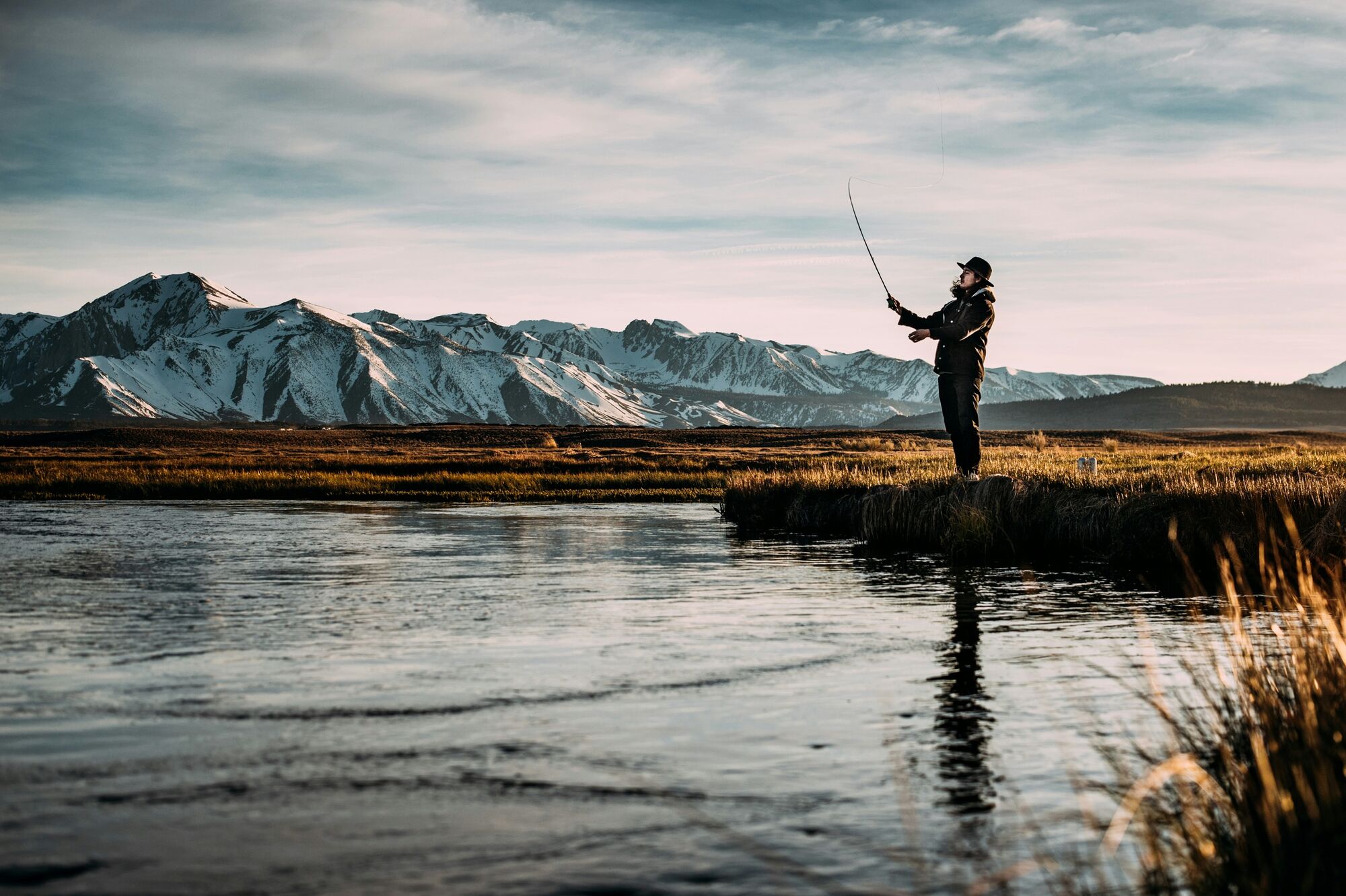 An angler casting a fly line from a grassy river bank, snow-capped mountains behind.