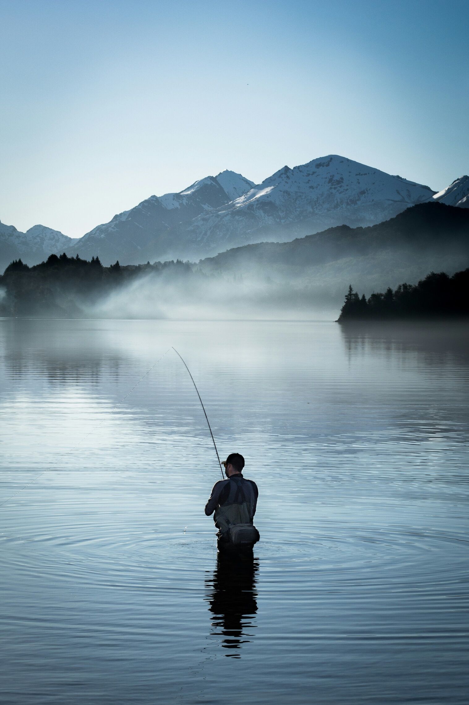 An angler wading in a misty mountain lake at first light.
